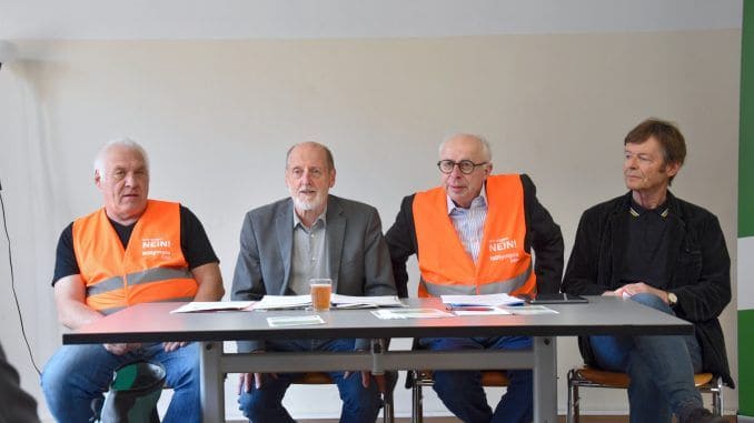 Vier Männer bei einer Diskussion, zwei tragen orange Warnwesten, im Hintergrund eine weiße Wand.