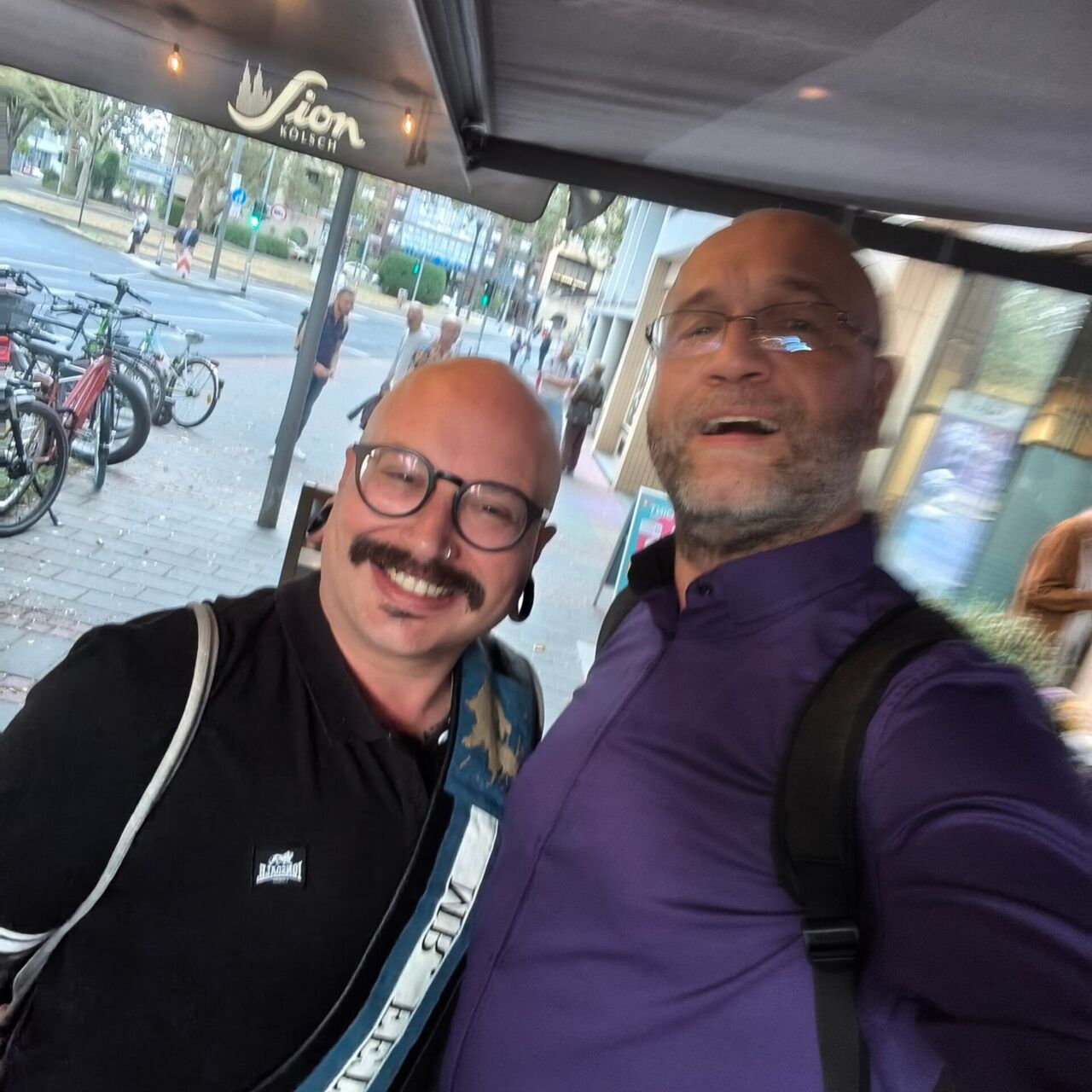 Zwei Männer lächeln beim Treffen im Café, draußen mit Fahrrädern im Hintergrund.