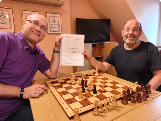 Zwei Männer spielen Schach, einer übergibt Dokument, im Hintergrund Fernseher.