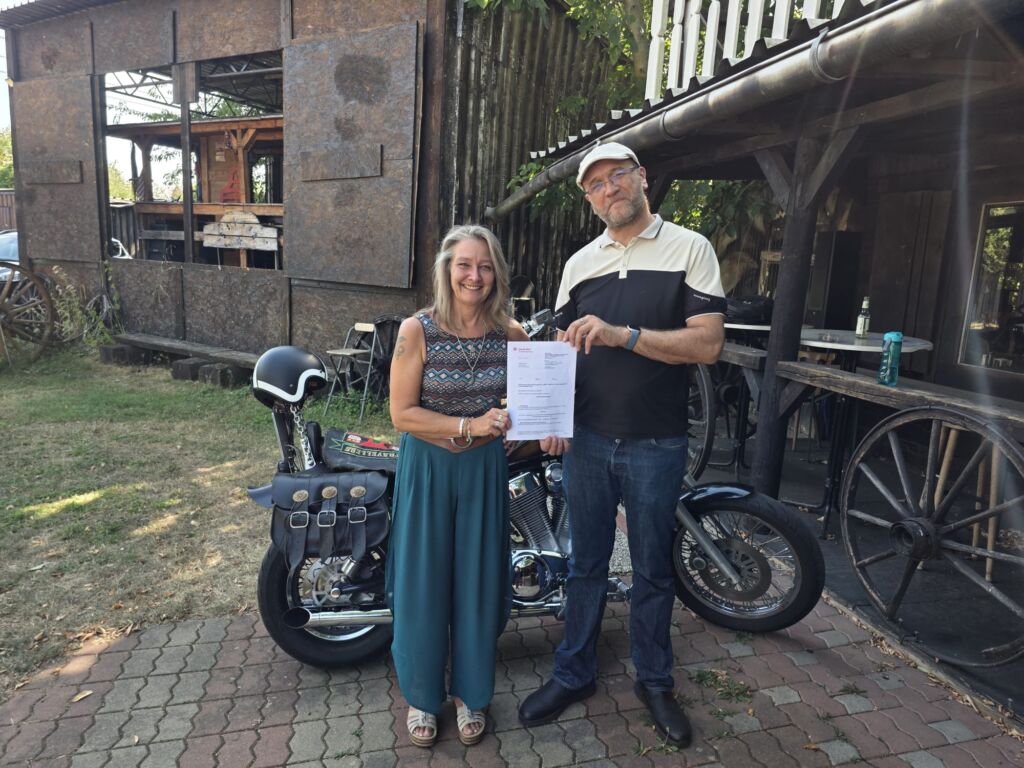 Dirk Bachhausen übergibt Dokument an Frau vor Motorrad im Garten.