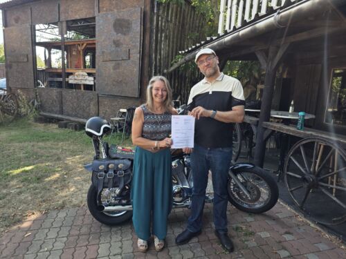 Dirk Bachhausen übergibt Dokument an Frau vor Motorrad im Garten.