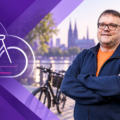 Dirk Bachhausen vor Kölns Skyline und Fahrrädern am Fluss.