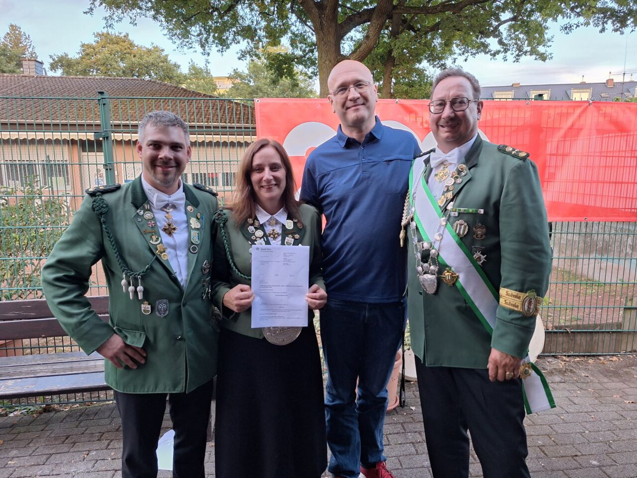 Vier Personen, darunter zwei in grünen Uniformen, bei Zeremonie im Freien.