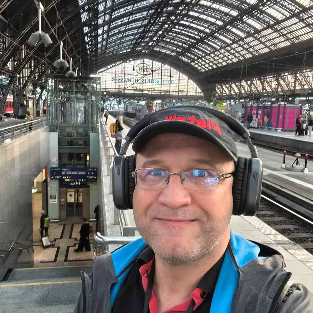 Mann mit Kopfhörern, Brille und Cap am Bahnhof, fotografiert im Selfie-Stil.