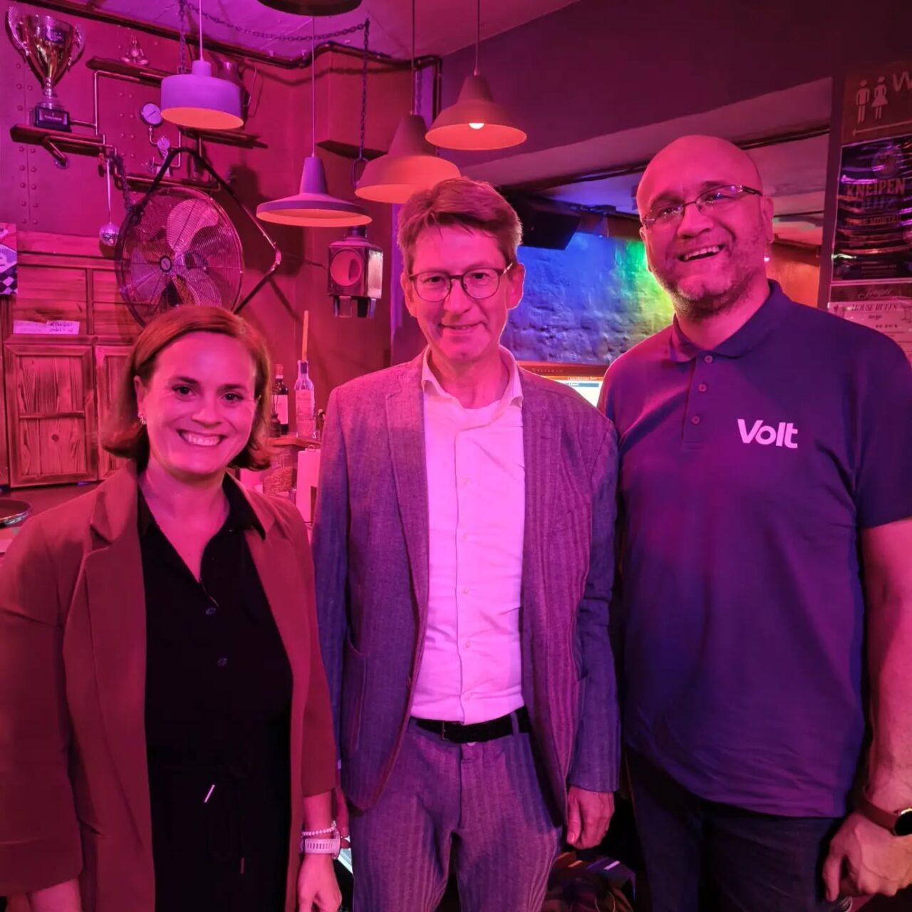 Drei Personen in einer Bar, lachend und freundlich, bunte Beleuchtung, entspannte Atmosphäre.